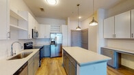 This well-designed single-floor residence offers a cohesive blend of modern finishes and functional living spaces throughout. The open-concept kitchen serves as the heart of the home, anchored by a large central island with a quartz countertop ideal for dining or food preparation. Cabinetry features a striking two-tone design — white upper cabinets paired with gray lower cabinets — complemented by a white subway tile backsplash and a full suite of stainless steel appliances, including a French door refrigerator with ice and water dispenser, electric range, microwave, and dishwasher. Brushed gold pendant lights hang above the island, adding a refined touch. The kitchen flows seamlessly into the living area, which is finished with warm hardwood-style flooring and ceiling fans, and opens to the outdoors through glass French doors that invite natural light. A carpeted bedroom provides a softer, quieter retreat, also benefiting from natural light through sizable windows with views of surrounding greenery. The bathroom is appointed with granite countertops, gray shaker-style cabinetry, a large vanity mirror, and a walk-in shower. Throughout the residence, neutral tones, clean lines, and quality materials create a contemporary and comfortable living environment.