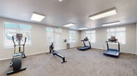 Located at 1030 S Chelton Rd in Colorado Springs, Colorado, this fitness center offers a well-appointed, single-floor exercise facility designed to support a range of workout routines. The space is bright and airy, with multiple windows fitted with horizontal blinds lining the walls and allowing generous natural light to fill the room throughout the day. Overhead, a series of recessed flat-panel light fixtures provide consistent, even illumination across the entire floor area.

The fitness center is equipped with a variety of cardio machines, including a treadmill, elliptical trainer, rowing machine, and an additional cardio unit, giving users diverse options for aerobic exercise. A folding exercise mat is also available, stored upright in a dedicated corner area, providing flexibility for floor-based stretching or bodywork. The flooring throughout consists of durable, low-pile carpet tile in a neutral tone, well-suited for a fitness environment.

The layout is open and spacious, offering ample room to move freely between equipment without feeling crowded. Neutral-toned walls and a clean, modern aesthetic contribute to an inviting and functional atmosphere. Two access doors provide convenient entry and exit points. A small waste receptacle and sanitation station near the entrance reflect attention to cleanliness and user comfort. Overall, the space is practical, well-lit, and thoughtfully organized for everyday fitness use.