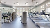 Located at 8801 Citrus Village Dr in Tampa, Florida, this fitness center offers a well-equipped exercise room designed to support a wide range of workout routines. The space features a clean, bright aesthetic with white walls, recessed and flush-panel lighting, and durable rubber flooring suited for high-traffic athletic use. Large multi-pane windows along the walls invite generous natural light and provide views of the surrounding greenery, creating an open and energizing atmosphere. Ceiling fans contribute to comfortable air circulation throughout the room. Full-length mirrors line multiple walls, visually expanding the space and allowing users to monitor their form during exercise. The cardio section is stocked with a row of treadmills and additional aerobic machines, while the strength training area includes a multi-station cable machine, a lat pulldown apparatus, weight benches, and a well-organized dumbbell rack with a complete range of free weights. Step platforms and other functional training accessories round out the equipment selection. A set of French doors with transom windows opens to the exterior, enhancing the connection to the outdoors. Fitness center rules are posted near the entrance, reflecting an organized and community-focused facility.