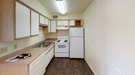 This single-floor unit offers a practical and well-organized layout designed to maximize livability. The kitchen is efficiently arranged in an L-shaped configuration, featuring white cabinetry with light wood trim, a double-basin sink, coil-top electric range, full-size refrigerator, and laminate countertops in a warm neutral tone. Dark wood-style vinyl flooring runs throughout the kitchen, adding contrast to the light-colored walls and cabinetry.

The living area is generously sized, with notable vaulted ceilings that lend a sense of openness and airiness to the space. Plush wall-to-wall carpeting in a warm beige tone covers the floor, contributing to a comfortable atmosphere. A large window along one wall allows natural light to flow in, while a glass-paneled exterior door provides direct outdoor access and additional light. A six-panel interior entry door anchors the front of the space.

The bedroom is well-proportioned, also finished with soft carpeting and featuring direct access to a private en-suite bathroom. The bathroom includes a single-sink vanity with storage drawers, light wood cabinetry, a wall-mounted mirror, and a tiled shower area. Dark vinyl flooring continues from the kitchen into the bathroom, maintaining a consistent aesthetic. The overall unit presents a clean, neutral palette suited for comfortable everyday living.