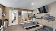 This dedicated fitness center offers a comprehensive workout facility designed to support a wide range of exercise routines. The space is finished with warm taupe-toned walls, carpeted flooring in the cardio area, and rubber mat flooring beneath the weight equipment, providing both comfort and functionality.

The cardio zone is equipped with multiple Life Fitness treadmills and elliptical trainers, positioned to face wall-mounted flat-screen televisions for an engaging workout experience. A recumbent stationary bike provides an additional low-impact option. Large windows with white blinds line the walls, allowing ample natural light to fill the room while offering views of the surrounding greenery.

The strength training area features a large multi-station cable weight machine capable of supporting a full-body resistance workout, complemented by a dedicated weight bench and free weights. A floor-length mirror spans one wall, enhancing the sense of space and allowing for proper form checks during exercise.

The room is accessible via a glass-paneled door and includes recessed ceiling lighting for consistent illumination. Posted facility rules ensure a well-managed environment. The overall ambiance is bright, clean, and purpose-built, making this fitness center a practical and fully appointed amenity.