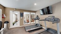 This dedicated fitness center offers a comprehensive workout facility designed to support a wide range of exercise routines. The space is finished with warm taupe-toned walls, carpeted flooring in the cardio area, and rubber mat flooring beneath the weight equipment, providing both comfort and functionality.

The cardio zone is equipped with multiple Life Fitness treadmills and elliptical trainers, positioned to face wall-mounted flat-screen televisions for an engaging workout experience. A recumbent stationary bike provides an additional low-impact option. Large windows with white blinds line the walls, allowing ample natural light to fill the room while offering views of the surrounding greenery.

The strength training area features a large multi-station cable weight machine capable of supporting a full-body resistance workout, complemented by a dedicated weight bench and free weights. A floor-length mirror spans one wall, enhancing the sense of space and allowing for proper form checks during exercise.

The room is accessible via a glass-paneled door and includes recessed ceiling lighting for consistent illumination. Posted facility rules ensure a well-managed environment. The overall ambiance is bright, clean, and purpose-built, making this fitness center a practical and fully appointed amenity.