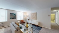 This single-floor residence offers a thoughtfully arranged layout that balances comfort and functionality across its key living spaces. The living room serves as the centerpiece of the home, featuring soft neutral walls, plush carpeting, and multiple windows dressed with white horizontal blinds that invite generous natural light. The space is well-proportioned, accommodating a full seating arrangement alongside a low-profile media console and decorative wall art, creating a warm and stylish atmosphere.

The kitchen is outfitted with warm oak cabinetry, granite-style countertops, and light hardwood-style flooring. It includes a full suite of appliances—refrigerator, dishwasher, and range—alongside ample cabinet and counter space arranged in a practical U-shaped configuration.

Two bedrooms provide comfortable accommodations. The primary bedroom is generously sized with carpet flooring, a large window, and room for a full dresser arrangement. The secondary bedroom features a soft sage accent wall, carpet flooring, a bright window, and space for a desk or sitting area, lending it versatility.

The bathroom presents a clean, functional design with gray tile surrounding a full soaking tub and shower combination, a single-sink vanity with cabinet storage below, and a large mirror. The overall ambiance throughout the residence is calm, neutral, and move-in ready.