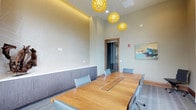 This single-floor conference room is thoughtfully designed to balance functionality with contemporary aesthetics. The centerpiece is a substantial warm-toned wood conference table with integrated cable management, surrounded by ergonomic gray upholstered chairs that comfortably accommodate multiple attendees. Three striking amber globe pendant lights cascade from the soaring ceiling, casting a warm, inviting glow throughout the room and serving as a bold design focal point.

The space features two distinct wall treatments: one wall is adorned with a subtle geometric patterned wallcovering above a run of sleek, low-profile gray cabinetry, providing both visual interest and practical storage. On the opposite side, large arched and rectangular windows with sheer white drapes allow abundant natural light to fill the room while offering views of the surrounding exterior landscape.

Warm wood baseboards and door trim complement the natural tones of the conference table, while patterned carpeting in a soft blue-gray palette adds texture underfoot. Curated artwork and a sculptural driftwood accent piece contribute to the polished, professional ambiance. A wood-framed glass door provides entry and maintains a sense of openness while delineating the space.