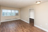 This single-floor unit offers a practical and comfortable layout with consistent hardwood-style flooring flowing throughout the main living areas and bedroom. The spacious living room is a standout feature, characterized by large windows dressed with vertical blinds that allow generous natural light to fill the space. A wall-mounted air conditioning unit and baseboard heating ensure year-round climate comfort. Neutral beige tones on the walls and a textured ceiling contribute to the room's clean, understated aesthetic. A doorway leads from the living area into the bedroom, which mirrors the warm flooring and features its own window with vertical blinds and baseboard heating, maintaining a bright and cozy atmosphere. The galley-style kitchen is efficiently designed with rich, dark wood cabinetry lining both sides, laminate countertops, a stainless steel sink, a white range, and a full-size refrigerator. Light tile flooring in the kitchen provides a practical contrast to the rest of the unit. The bathroom is neatly appointed with tile flooring, a vanity featuring matching dark wood cabinetry with a light stone-style countertop, a large mirror, and a full tub-shower combination with tile surround. Overall, the unit presents a functional and move-in-ready living space.