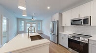 Located on Nexus Ave in Stafford, TX, this thoughtfully designed one-bedroom, one-bathroom residence offers a clean, modern aesthetic across a single floor. The open-concept kitchen and living area serves as the heart of the home, showcasing white shaker-style cabinetry, light gray lower cabinets, white quartz countertops, a subway tile backsplash, and a full suite of stainless steel appliances including a French door refrigerator, electric range, built-in microwave, and dishwasher. A center island with a deep farmhouse sink and pendant lighting above adds both functionality and visual appeal. The living area is bright and airy, benefiting from large windows and glass French doors that invite natural light and provide outdoor views, complemented by a ceiling fan overhead. Warm wood-look flooring flows throughout the main living spaces, while the bedroom features plush carpeting, a ceiling fan, and a window overlooking the exterior. The bathroom is finished with marble-style tile surrounding the tub and shower, a wide white vanity with an illuminated mirror, and matching wood-look flooring. An in-unit stacked washer and dryer and a dedicated closet round out the practical offerings of this well-appointed residence.