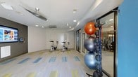 This fitness room offers a clean, functional space designed to support a variety of workout routines. The room features a neutral color palette with white walls and light gray tones that contribute to a bright, open atmosphere. Recessed lighting is complemented by ceiling fans, ensuring the space remains well-lit and comfortable during exercise. The flooring consists of a patterned carpet tile in muted blues, greens, and yellows, adding visual interest while providing a durable surface underfoot. Stationary cycling bikes are positioned toward the rear of the room, offering a dedicated cardio station. Along one side, a vertical storage rack holds multiple stability balls in varying sizes and colors, alongside resistance bands and additional accessories, keeping equipment organized and accessible. A mirrored wall panel near the equipment area enhances the sense of space and allows users to monitor their form. Glass-panel doors connect the fitness area to an adjacent space, maintaining an open, airy feel throughout. The single-story layout keeps everything conveniently accessible, making this a practical and well-appointed fitness facility.