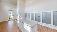 Located at 8215 Dougherty Ct in Travelers Rest, South Carolina, this single-story mail center is designed to serve a residential community with a practical yet polished aesthetic. The space immediately impresses with its dramatically vaulted ceilings, which lend an airy, open feel that extends well beyond what its footprint might suggest. Crisp white walls and an abundance of natural light, drawn in through high clerestory windows and a glass-paneled entry door, create a bright and welcoming environment throughout the day.

The flooring consists of warm wood-look planks that run continuously through the space, complementing the clean, neutral palette of the interior. A central island finished with a granite-patterned countertop provides a convenient surface for sorting and managing mail. Along the walls, multiple banks of individual steel mailboxes are neatly organized and accessible, accommodating a large number of residents. A dedicated parcel compartment system is also integrated into the mailbox units for package delivery.

The entry area features a glass door framed by stone exterior cladding, offering a clear sightline to the outdoors. Safety amenities, including a mounted fire extinguisher and illuminated exit signage, are prominently positioned. Overall, the space balances function and design with a clean, modern finish.