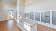 Located at 8215 Dougherty Ct in Travelers Rest, South Carolina, this single-story mail center is designed to serve a residential community with a practical yet polished aesthetic. The space immediately impresses with its dramatically vaulted ceilings, which lend an airy, open feel that extends well beyond what its footprint might suggest. Crisp white walls and an abundance of natural light, drawn in through high clerestory windows and a glass-paneled entry door, create a bright and welcoming environment throughout the day.

The flooring consists of warm wood-look planks that run continuously through the space, complementing the clean, neutral palette of the interior. A central island finished with a granite-patterned countertop provides a convenient surface for sorting and managing mail. Along the walls, multiple banks of individual steel mailboxes are neatly organized and accessible, accommodating a large number of residents. A dedicated parcel compartment system is also integrated into the mailbox units for package delivery.

The entry area features a glass door framed by stone exterior cladding, offering a clear sightline to the outdoors. Safety amenities, including a mounted fire extinguisher and illuminated exit signage, are prominently positioned. Overall, the space balances function and design with a clean, modern finish.