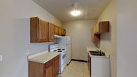This single-floor residential space is centered around a well-organized galley-style kitchen that makes efficient use of its layout. The kitchen features warm oak-toned wood cabinetry both above and below the countertops, providing ample storage for everyday essentials. Upper cabinets line both sides of the kitchen, maximizing vertical storage space, while lower cabinets offer additional room beneath light-colored countertops. White appliances, including a coil-top electric range and a full-size refrigerator, are neatly integrated into the layout. A range hood above the stove provides practical ventilation. On the opposite wall, a double-basin sink with a chrome faucet sits beneath additional cabinetry and open shelving, offering a convenient prep and cleanup area. The flooring throughout the kitchen consists of classic patterned vinyl tiles in neutral tones, contributing to the clean and understated aesthetic. A textured ceiling adds a subtle design element overhead, and flush-mount ceiling lighting ensures the space is well lit. The overall ambiance of the kitchen is practical and functional, with a timeless, homey character defined by its consistent wood tones and straightforward design.