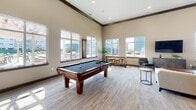 Located at 4131 W 54th St N in Sioux Falls, South Dakota, this single-floor amenity facility at Pinnacle Point is designed to offer a well-rounded communal experience. The space is thoughtfully organized into several distinct functional areas, each finished with neutral tones, plush carpeting, and consistent modern detailing throughout.

The entrance foyer greets visitors with glass-paneled doors bearing the Pinnacle Point branding, alongside a glass-enclosed office or leasing area equipped with workstations and seating. Double glass doors with dark wood frames open into the main interior corridor, leading to the primary amenity spaces.

The expansive lounge features soaring ceilings with crown molding, abundant natural light from large windows overlooking the surrounding development, and a comfortable seating arrangement centered around a wall-mounted flat-screen television. Adjacent to the lounge, a game room mirrors the same generous ceiling height and natural light, highlighted by a full-size billiards table and casual seating areas.

A fully equipped kitchen includes dark shaker-style cabinetry, quartz countertops, stainless steel appliances, and a multi-seat island, accompanied by a nearby dining area. The fitness center occupies a dedicated wing, offering cardio machines, free weights, wall-mounted televisions, and natural light from exterior-facing windows.
