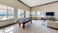 Located at 4131 W 54th St N in Sioux Falls, South Dakota, this single-floor amenity facility at Pinnacle Point is designed to offer a well-rounded communal experience. The space is thoughtfully organized into several distinct functional areas, each finished with neutral tones, plush carpeting, and consistent modern detailing throughout.

The entrance foyer greets visitors with glass-paneled doors bearing the Pinnacle Point branding, alongside a glass-enclosed office or leasing area equipped with workstations and seating. Double glass doors with dark wood frames open into the main interior corridor, leading to the primary amenity spaces.

The expansive lounge features soaring ceilings with crown molding, abundant natural light from large windows overlooking the surrounding development, and a comfortable seating arrangement centered around a wall-mounted flat-screen television. Adjacent to the lounge, a game room mirrors the same generous ceiling height and natural light, highlighted by a full-size billiards table and casual seating areas.

A fully equipped kitchen includes dark shaker-style cabinetry, quartz countertops, stainless steel appliances, and a multi-seat island, accompanied by a nearby dining area. The fitness center occupies a dedicated wing, offering cardio machines, free weights, wall-mounted televisions, and natural light from exterior-facing windows.
