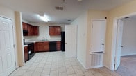 This single-floor space presents a practical and straightforward layout centered around a well-equipped kitchen. The kitchen features warm-toned wooden cabinetry that lines the walls, providing ample storage for everyday essentials. Black appliances, including a refrigerator, dishwasher, and range, offer a cohesive contrast against the lighter elements of the room. A built-in sink is positioned along the counter, adding to the kitchen's functional design.

The flooring throughout the space consists of light-colored ceramic tile, which contributes to a clean and cohesive look while being easy to maintain. The walls are finished in a neutral off-white tone, creating a bright and open atmosphere. Overhead fluorescent lighting ensures the kitchen area is well-lit for daily tasks.

Ventilation is provided through ceiling-mounted HVAC vents, and a floor-level register near the entryway ensures consistent climate control throughout the space. Two interior doors are visible, suggesting additional rooms or storage areas beyond the main open area. Overall, the space offers a no-frills, functional environment suitable for everyday living.
