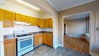This 2-bedroom, 1-bathroom unit at 600 James St in Syracuse, NY offers a well-structured single-floor layout with classic character throughout. The kitchen is efficiently arranged in an L-shape, featuring warm honey-toned oak cabinetry, dark granite-look countertops, a gas range, refrigerator, and under-cabinet range hood. An arched entryway adds an architectural charm, connecting the kitchen to the rest of the unit. The flooring transitions from durable vinyl tile in the kitchen to rich hardwood floors that extend through the living areas and bedrooms. The living room is generously proportioned, showcasing panel-detailed walls, crown molding, a ceiling fan, and multiple windows that bathe the space in natural light. Both bedrooms are well-sized, each featuring hardwood floors, large windows with views of the surrounding neighborhood, and radiator heating units. One bedroom also includes a closet. The bathroom is equipped with a full soaking tub, a single vanity with sink, and track lighting. Throughout the unit, neutral taupe wall tones provide a calm, versatile backdrop, while the abundance of natural light and classic architectural details lend the space a timeless residential character.