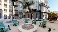 This contemporary residential amenity space is thoughtfully designed to foster community and comfort across multiple well-appointed areas. The welcoming entry features floor-to-ceiling glass double doors that flood the space with natural light and offer clear views to the surrounding landscaping and parking area, complemented by a warm natural wood reception desk.

The outdoor courtyard serves as a central gathering hub, anchored by a circular stone fire pit surrounded by green Adirondack chairs. Tall palm trees rise above the space, adding a relaxed, resort-like atmosphere, while a spa or water feature and picnic seating areas provide additional lounging options—all framed by the multi-story building facade finished in contrasting grey stone and dark wood trim.

Inside, a fully equipped communal kitchen showcases light wood cabinetry, stainless steel appliances, sleek countertops, and statement geometric pendant lighting, creating an ideal setting for social gatherings. Adjacent to the kitchen, a spacious co-working lounge features long communal tables, mixed seating, industrial-style stools, and a glass garage-style door that opens the room to fresh air and natural light. A decorative wall-mounted art piece adds character to the lounge area. Adding a playful touch, one interior room features charming cactus-patterned accent walls, lending a whimsical, Southwest-inspired personality to the space.
