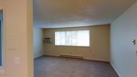 This single-floor space features a spacious main living area finished with wall-to-wall carpeting in a neutral tone, contributing to a quiet and comfortable atmosphere. The room is illuminated by a large multi-pane window fitted with horizontal blinds, allowing a generous amount of natural light to fill the interior while offering adjustable privacy. The walls are painted in a soft, warm beige, lending the space a clean and understated aesthetic. Climate control is well-addressed, with a wall-mounted air conditioning unit on one side of the room and a baseboard heating system running along the exterior wall beneath the window, ensuring year-round comfort. The ceiling features a textured popcorn finish, a characteristic detail of the space's construction. Trim and baseboards run along the perimeter, providing a tidy, finished look throughout. The overall layout is open and unobstructed, offering flexibility in how the space can be arranged and utilized. The neutral palette and practical fixtures make this a functional and adaptable living environment.