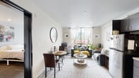 This single-floor residence offers a thoughtfully designed layout that balances comfort and functionality. The open-concept living and dining area is bathed in natural light from a large window dressed with sheer white curtains, offering pleasant outdoor views. Neutral tones on the walls and a patterned area rug create a clean, welcoming ambiance, while a round mirror accent adds a touch of style to the dining nook. A compact yet fully equipped kitchen lines one wall, showcasing dark cabinetry with open upper shelving, granite-style countertops, and a complete suite of stainless steel appliances including a refrigerator, range, microwave, and dishwasher. The kitchen flows seamlessly into the living space, making it ideal for everyday use. The bedroom is a serene retreat, featuring large vertical-blind windows that allow generous natural light and views of greenery outside. Carpeted flooring and ample space accommodate a full sleeping and workspace setup comfortably. The bathroom is finished with granite countertops and neutral tiling, providing a clean and functional environment. Throughout the residence, recessed lighting and crown molding details contribute to a polished, modern aesthetic.