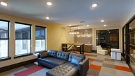 This single-floor space offers a well-designed, open-concept layout that seamlessly blends comfort and modern aesthetics. The lounge area anchors the space with a large tufted leather sectional sofa positioned atop a vibrant geometric-patterned carpet featuring bold red, orange, and gray tones, adding a dynamic visual focal point to the room.

Large windows along the perimeter flood the space with natural light, creating a bright and welcoming atmosphere. The warm yellow walls complement the contemporary design, while recessed lighting throughout ensures a well-lit environment at any time of day.

A bar-height communal table with industrial-style metal stools serves as a casual dining or collaborative work area, positioned beneath a striking Edison bulb pendant light fixture that adds an industrial-chic touch. Behind it, a tiled accent wall frames built-in cabinetry, lending a refined, finished look to the space.

An adjacent seating nook with white upholstered chairs offers a quieter retreat within the open layout, providing versatility for various uses. The overall ambiance is modern and energetic, making this space well-suited for socializing, working, or unwinding in equal measure.