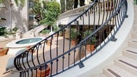 Located in the heart of downtown Los Angeles, this distinguished outdoor space at 725 S Bixel St showcases a refined Mediterranean-inspired design that blends classical architectural elegance with lush landscaping. A sweeping curved staircase serves as a central architectural focal point, finished in terrazzo-style stone with rich wood inlay accents on each step and embedded low-profile lighting for ambiance and safety. The staircase is flanked by ornate wrought iron railings featuring scrollwork detailing, lending a timeless, sophisticated character throughout.

The expansive terrace level opens to a generously proportioned open-air promenade finished in warm stone tile, lined with terracotta planters overflowing with palms, cypress trees, tropical foliage, and seasonal flowering plants. Classical stone balustrades border the perimeter, reinforcing the space's stately aesthetic while framing panoramic views of the Los Angeles downtown skyline and surrounding urban canopy. A central courtyard below features a decorative fountain and additional greenery, contributing to a serene, resort-like atmosphere. The abundant natural light and open sky above enhance the airy, inviting quality of this meticulously curated outdoor environment.