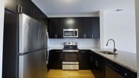 The kitchen is thoughtfully designed with a bold contrast between dark espresso-toned cabinetry and light-colored walls, creating a striking visual dynamic. Upper and lower cabinets with brushed metal hardware offer generous storage throughout the space. Stainless steel appliances, including a French door refrigerator, an electric range with oven, and an over-the-range microwave, provide a cohesive, modern aesthetic. The countertops feature speckled granite in neutral tones, offering both durability and style. A subway tile backsplash in white adds a clean, classic touch between the upper and lower cabinetry. The kitchen is arranged in a U-shaped layout, maximizing workspace efficiency and ease of movement. A stainless steel sink is positioned along the perimeter, complemented by a modern gooseneck faucet. Warm hardwood flooring grounds the space and softens the contrast of the dark cabinetry above. Recessed overhead lighting ensures the kitchen remains bright and functional. Overall, the space balances modern design sensibilities with practical, well-organized functionality.