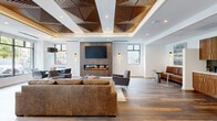 Located at 22 Schuyler Place in Morristown, NJ, this thoughtfully designed lounge space delivers a refined contemporary aesthetic across a single open floor. The centerpiece of the living area is a dramatic wood-paneled accent wall housing a wide linear electric fireplace and a wall-mounted television, creating a warm yet modern focal point. Above, an intricate geometric coffered ceiling with interlocking wood slat panels adds architectural depth and visual interest, complemented by recessed lighting throughout.

Wide-plank wood-look flooring runs continuously across the space, grounding the open layout with warmth and texture. Multiple windows along the perimeter invite abundant natural light and views of the surrounding streetscape. The lounge area is furnished with a generous leather sofa, accent chairs, and a glass coffee table, while a separate dining area accommodates round-table seating nearby.

A secondary nook along one wall features built-in leather banquette seating alongside a compact wet bar with warm wood cabinetry, mosaic tile backsplash, and gold hardware — ideal for casual gatherings. Bold steel-framed glass partition walls and doors define the entry corridor, lending an industrial-chic transparency to the overall design. The result is a polished, versatile communal space with a welcoming and upscale ambiance.