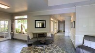 This single-floor space presents a welcoming and polished interior designed for both comfort and functionality. The main seating area features a contemporary gray sofa accented with decorative pillows, centered around a sleek oval coffee table resting on a geometric-patterned area rug. A framed mirror with a bold black border anchors the wall above, adding depth and visual interest to the space.

The flooring throughout is composed of neutral-toned large-format tiles, contributing to a clean and cohesive aesthetic. Crown molding runs along the upper walls, lending a refined architectural detail to the interior. The color palette is kept light and airy, with soft white and gray tones creating a calm, professional ambiance.

Large windows along the exterior wall flood the seating area with natural light and offer views of lush palm trees and greenery outside. French-style glass doors with transom windows lead into an extended corridor, which is lined with wall-mounted fixtures and additional framed artwork. The hallway stretches generously, maintaining the same bright, open character as the main area. Overall, the space balances modern furnishings with classic architectural details to create an inviting and informative environment.