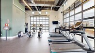 Located in Grove City, Ohio, this single-floor fitness center offers a well-appointed exercise space designed for a comprehensive workout experience. The facility is defined by its striking architectural elements, including soaring ceilings with an exposed industrial-style framework, linear LED lighting, and oversized glass-panel garage doors that flood the interior with natural light and provide open views of the outdoor surroundings.

The main exercise room features durable rubber flooring throughout, providing both safety and functionality. Cardio equipment is prominently represented with multiple treadmills and elliptical machines arranged to take full advantage of the natural light from the large glass doors. A dedicated strength and functional training area includes a multi-station cable machine, an adjustable weight bench, and a well-stocked rack of medicine balls, resistance tools, and exercise accessories. Wall-mounted televisions are positioned throughout for entertainment during workouts.

A secondary area, separated by sleek glass-and-steel partition walls, provides a more intimate space for stretching, cycling, or focused training. The entry corridor features glass-panel doors with posted fitness policies, leading into the main workout floor. Accent walls in muted sage green and slate blue add a modern, welcoming aesthetic. The overall ambiance is bright, open, and energizing, making this fitness center a fully functional and visually appealing space.