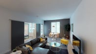 This single-floor residence showcases a thoughtfully designed layout that balances open communal spaces with private quarters. The living area serves as the heart of the unit, featuring neutral-toned walls contrasted by a bold charcoal accent wall, generous natural light from large windows with horizontal blinds, and room for both a sectional seating arrangement and a dedicated dining area. A sliding barn-style door connects the living space to the bedroom, adding a modern architectural detail while maximizing flow.

The kitchen is well-appointed with dark espresso cabinetry, stainless steel appliances including a refrigerator, range, and built-in microwave, complemented by a subway tile backsplash and a light quartz peninsula countertop with pendant lighting above. The peninsula provides additional seating and prep space, making it a functional centerpiece.

The bedroom is a calm retreat with soft neutral tones, natural light from a window with blinds, and ample space for furnishings. The bathroom features an elongated vanity with dual sinks, stone-look countertops, dark cabinetry, and a tub-shower combination. A stacked in-unit washer and dryer adds everyday convenience. Throughout, pops of yellow and teal infuse the space with a lively, contemporary character.