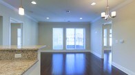 This single-floor space showcases a thoughtfully designed open layout that seamlessly connects the kitchen and living areas. Dark hardwood floors run throughout, adding warmth and a polished aesthetic to the interior. Crown molding lines the ceiling, lending a refined architectural detail that complements the neutral, light-toned walls.

The kitchen features a granite-topped breakfast bar, providing both a functional workspace and a natural divider between the cooking and living zones. Recessed lighting is installed throughout the ceiling, ensuring even, ambient illumination across the space. A ceiling-mounted light fixture adds an additional design element near the kitchen area.

The living area is bright and airy, benefiting from natural light streaming through a set of windows fitted with horizontal blinds. A sliding glass door opens to an outdoor balcony or patio, offering exterior views and easy access to fresh air. The combination of recessed lighting, hardwood flooring, crown molding, and granite surfaces gives the space a clean, contemporary feel with classic finishing touches throughout.