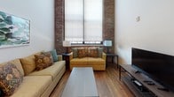 Located at 220 Merrimack in Lawrence, MA, this single-floor unit masterfully blends industrial heritage with modern finishes. Warm hardwood floors and soaring ceilings run throughout, creating an open, airy atmosphere. Exposed brick accent walls add authentic character to both the living area and bedrooms, while large windows flood the space with natural light and street views.

The kitchen is outfitted with dark cabinetry, stainless steel appliances including a full-size refrigerator, range, and built-in microwave, complemented by white quartz countertops, a subway tile backsplash, and open wooden shelving. A peninsula with seating bridges the kitchen and dining area seamlessly.

The living room features soaring ceilings, exposed brick flanking oversized windows, and rich hardwood flooring, creating a welcoming and spacious gathering area. Both bedrooms are generously proportioned with high ceilings and walk-in closets. One bedroom showcases exposed brick and large street-facing windows, while the other offers a quieter, refined atmosphere.

The bathroom is elegantly appointed with white subway tile walls, hexagonal floor tile, a walk-in glass shower with gold hardware, a modern vessel sink, and a warm wood vanity, delivering a crisp, upscale finish throughout.