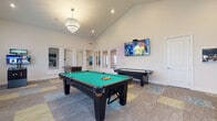 Located at 484 Greenmont Dr in Lewis Center, Ohio, this single-floor amenity facility is thoughtfully designed for recreation and social engagement. The centerpiece is a spacious game room boasting soaring vaulted ceilings accented by an elegant crystal chandelier. The room is equipped with a full-size billiards table, an air hockey table, a foosball table, and a wall-mounted flat-screen television, all set atop vibrant multi-toned patterned carpet tiles that add a lively energy to the space.

Adjacent to the game room is the "OG Café" lounge area, where a neon-lit accent wall in deep navy blue anchors a stylish café-style bar counter with marble-finish surfaces and pendant lighting. Round tables with contemporary tulip-style chairs offer casual seating, while a fitness center is visible through glass partitions, keeping wellness close at hand.

Two dedicated poker rooms provide a more intimate setting, each featuring a full-size poker table with leather seating, wall-mounted televisions, and large windows that invite natural light and views of the surrounding neighborhood. Sliding barn doors offer flexible privacy between spaces. Thoughtful décor throughout, including curated artwork and lush tropical plants, adds character and warmth to this well-appointed community retreat.