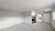 This single-floor unit is designed with a clean, neutral aesthetic throughout, featuring light grey walls and plush grey carpeting across the main living and bedroom areas. The open layout connects the living space to the kitchen, creating a functional flow between rooms.

The spacious living area is equipped with a ceiling fan and benefits from natural light through multiple windows fitted with blinds. Baseboard heating runs along the exterior walls, complemented by a wall-mounted air conditioning unit for year-round climate control. The entry area features a double-door setup providing both a main entrance and a closet, along with a structural column detail that adds definition to the space.

The kitchen is well-appointed with white shaker-style cabinetry, granite-look countertops, a coordinating tile backsplash, a stainless steel sink, and a stainless steel gas range with an overhead ventilation hood. Adjacent to the kitchen, a separate bedroom offers carpeted flooring, a window with natural light, and baseboard heating.

The bathroom presents a clean, all-white design featuring a full tub-shower combination and wood-look vinyl flooring. Throughout the unit, the consistent grey-and-white palette creates a cohesive, move-in-ready atmosphere.