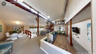 The Paddington Court Clubhouse in Raleigh, NC is a well-appointed community gathering space defined by its striking architectural character and versatile layout. Soaring vaulted ceilings and bold exposed dark-wood timber beams and posts create a dramatic, lodge-inspired aesthetic throughout the main floor. Pendant globe lights and ceiling fans complement the abundant natural light that pours in through transom and full-height windows, lending the interior a bright and welcoming ambiance.

The open-plan layout is thoughtfully divided into distinct functional zones. A spacious lounge area features built-in seating with teal upholstery and green accent pillows alongside contemporary armchairs, all arranged to encourage comfortable gathering. Adjacent to the lounge, a dining area is outfitted with rich wood-topped tables and sage green chairs, with direct access to an outdoor deck through glass-paned French doors framed by colorful chevron-patterned valances. Views of surrounding wooded greenery enhance the natural atmosphere.

A full kitchen with white cabinetry and stainless steel appliances supports events and gatherings. A striking wood staircase with dark metal balusters leads to an open upper mezzanine level overlooking the main floor. The combination of hardwood-style flooring and plush carpet areas, curated artwork, and cohesive teal and green accents throughout gives the clubhouse a polished yet inviting character.