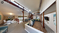The Paddington Court Clubhouse in Raleigh, NC is a well-appointed community gathering space defined by its striking architectural character and versatile layout. Soaring vaulted ceilings and bold exposed dark-wood timber beams and posts create a dramatic, lodge-inspired aesthetic throughout the main floor. Pendant globe lights and ceiling fans complement the abundant natural light that pours in through transom and full-height windows, lending the interior a bright and welcoming ambiance.

The open-plan layout is thoughtfully divided into distinct functional zones. A spacious lounge area features built-in seating with teal upholstery and green accent pillows alongside contemporary armchairs, all arranged to encourage comfortable gathering. Adjacent to the lounge, a dining area is outfitted with rich wood-topped tables and sage green chairs, with direct access to an outdoor deck through glass-paned French doors framed by colorful chevron-patterned valances. Views of surrounding wooded greenery enhance the natural atmosphere.

A full kitchen with white cabinetry and stainless steel appliances supports events and gatherings. A striking wood staircase with dark metal balusters leads to an open upper mezzanine level overlooking the main floor. The combination of hardwood-style flooring and plush carpet areas, curated artwork, and cohesive teal and green accents throughout gives the clubhouse a polished yet inviting character.