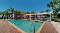 This outdoor amenity space offers a well-appointed, resort-inspired environment designed for relaxation and recreation. At the heart of the area sits a generously sized rectangular swimming pool with crystal-clear turquoise water, accessible via a classic metal entry rail and framed by a brick paver pool deck. Flanking one side of the pool is a striking white pergola structure, providing partial shade over a row of dark-toned lounge chairs — ideal for sunbathing while staying cool. The expansive brick paver courtyard extends beyond the pool area, offering ample open space that accommodates outdoor dining sets with umbrella-shaded tables and additional lounge seating. Lush tropical landscaping, including tall swaying palm trees and manicured greenery, lines the perimeter and enhances the warm, Florida-inspired atmosphere. Black ornamental iron fencing defines the boundaries of the space, adding a sense of enclosure and security while maintaining visibility to the surrounding grounds. The adjacent building features a coastal-style blue exterior with a pitched roof, complementing the overall aesthetic of the outdoor setting. Under abundant natural sunlight and open skies, this amenity area presents a vibrant and inviting environment well-suited for leisure and outdoor gatherings.