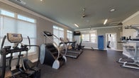 Located at 5090 Tradeway Dr in Leland, NC, this fitness center is a comprehensive, single-floor facility designed to support a full range of workout routines. The space is thoughtfully organized into distinct functional zones, all tied together by durable rubber flooring, neutral gray walls, and crisp crown molding details that lend a polished, well-maintained appearance throughout.

The cardio area is generously sized and equipped with multiple treadmills, elliptical machines, a stair climber, and a rowing machine. Large windows fitted with white blinds line the walls, allowing abundant natural light to fill the space, while ceiling fans help maintain a comfortable environment during workouts. Wall-mounted televisions provide additional entertainment options.

The strength training zone offers a well-stocked selection of dumbbells displayed on tiered racks, an adjustable weight bench, a power rack, and a cable machine, providing users with versatile free-weight and resistance training options. A water fountain station is conveniently located in this area for easy access during workouts.

A separate cycling studio features hardwood-style flooring, spin bikes, wall-mounted medicine balls, and stability balls, creating a bright and energetic space for group or individual cycling sessions. Large windows offer views of the surrounding community. The facility also includes a sanitation station, ensuring a clean and safe environment for all users.