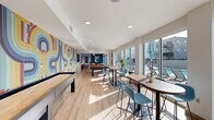 Located at 1050 W Colfax Ave in Denver, Colorado, this dynamic single-floor amenity space offers a generously sized, thoughtfully designed environment for socializing, working, and relaxing. The layout flows seamlessly across a living room and dining area, unified by light hardwood flooring and crisp white ceilings with recessed lighting throughout.

The living room is a standout feature, anchored by a bold teal slatted accent wall with a mounted flat-screen TV, vibrant orange lounge chairs, and a plush sofa arranged around a coffee table. Floor-to-ceiling windows flood the space with natural light and provide direct views of an outdoor terrace and pool area, with sliding glass doors offering seamless indoor-outdoor access.

Adjacent to the living area, a stylish wet bar with a teal island, white countertops, and blush bar stools serves as a social hub. A dramatic full-wall mural in colorful, retro-inspired patterns commands attention across the game area, which includes a shuffleboard table and pool table, adding an energetic, artistic focal point.

A private meeting room is finished with a warm terracotta accent wall, a wall-mounted display, a round wooden table, and modern pendant lighting—ideal for focused gatherings. The dining area mirrors this sophistication with curated artwork, high-top tables, and contemporary blue seating throughout.
