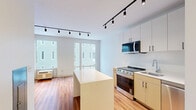 Located at 4240 Chestnut Street in Philadelphia's University City neighborhood, this single-floor studio offers a thoughtfully designed open-concept layout that combines the kitchen and living area into one cohesive space. Warm hardwood flooring runs throughout, complementing the bright, white walls and creating a clean, modern aesthetic.

The kitchen is well-appointed with a full suite of stainless steel appliances, including a range, over-the-range microwave, dishwasher, and refrigerator. Two-toned cabinetry — white uppers paired with warm tan lower cabinets — provides ample storage, while the white quartz countertops and a central island add both workspace and visual appeal. Black-hardware accents throughout lend a contemporary touch. Track lighting runs along the ceiling, offering targeted, adjustable illumination across the open living and kitchen zones.

The living area benefits from abundant natural light streaming through large windows and a floor-to-ceiling glass door, with views of the surrounding urban streetscape. The bathroom features a walk-in shower with white tile surround, a single vanity with a gray countertop, matching tan cabinetry, and continuous hardwood-style flooring. The overall design is minimal and modern, making efficient use of every square foot in this well-planned urban residence.