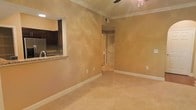 This single-floor residence offers a welcoming and functional layout designed with comfort in mind. The main living area is finished with plush wall-to-wall carpeting in a neutral tone, creating a soft and inviting atmosphere. Warm, earthy tones coat the walls throughout, contributing to a cozy and cohesive aesthetic. Crown molding runs along the ceiling perimeter, adding a touch of classic architectural detail, while a ceiling fan provides both style and practical air circulation.

A notable feature of the space is the pass-through opening that connects the main living area to the kitchen, enhancing the open feel and allowing for easy interaction between the two spaces. The kitchen is equipped with dark cabinetry and granite countertops, offering a rich contrast to the lighter tones of the living area.

A doorway leading to additional rooms — featuring louvered bifold doors — hints at further functional spaces such as a laundry closet or storage area. The interior is unified by clean white baseboards and trim that frame each room neatly. Overall, the space presents a straightforward, well-maintained layout with classic finishes suited for comfortable everyday living.