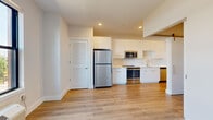 This thoughtfully designed single-floor unit offers a clean, modern aesthetic with an efficient layout. Light oak-toned hardwood flooring runs throughout the main living areas and hallway, providing warmth and visual continuity across the space. Recessed lighting and crisp white walls contribute to a bright, open atmosphere.

The kitchen is a standout feature, outfitted with sleek white flat-front cabinetry extending to the ceiling for ample storage. Stainless steel appliances — including a full-size refrigerator, range, microwave, and dishwasher — lend a professional, contemporary feel, while the white countertops and subway tile backsplash maintain a clean, cohesive look. A window at the end of the main room allows natural light to filter in, with views of the surrounding neighborhood.

The hallway connects the main living area to the bathroom and includes a utility panel integrated neatly into the wall. A walk-in closet provides practical storage. The bathroom is finished with light wood-grain cabinetry, a white undermount sink with clean countertops, a large mirror, and a matching wall-mounted storage cabinet. Light-toned tile flooring completes the serene, neutral palette. Overall, the unit presents a fresh, move-in-ready environment with modern finishes and efficient use of space.