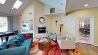 This single-floor clubhouse is a well-appointed community gathering space designed for comfort, recreation, and social interaction. The main lounge area immediately draws attention with its soaring vaulted ceilings that amplify the sense of openness and airiness. Warm yellow walls create an inviting atmosphere, while light hardwood flooring runs throughout, tying the spaces together cohesively. A marble-surround fireplace serves as the focal point of the lounge, complemented by a vibrant multicolored area rug, a teal sofa accented with red throw pillows, a carved wood coffee table, and a pair of neutral accent chairs. A decorative sunburst mirror above the fireplace adds a stylish finishing touch. French doors lead to an outdoor patio area, flooding the space with natural light and providing easy access to the exterior. The billiards room, separated by a partial wall, features vaulted ceilings, large windows with patterned drapery, and sweeping views of a pool and landscaped grounds — making it a naturally lit, recreational retreat. A dedicated refreshment area offers a countertop with appliances, cabinetry, and a bistro-style seating nook, ideal for casual meetings or a quick coffee break. Together, these spaces form a cohesive, lively clubhouse environment.
