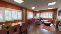 This single-floor space makes a strong visual impression with its bold, warm orange accent walls that flow throughout the main living and dining areas, creating an energetic and inviting atmosphere. Rich hardwood flooring runs continuously across the open-plan layout, tying together each distinct zone with a sense of cohesion and warmth.

The spacious living area is furnished with a sectional sofa, accent chairs in vibrant red, and a patterned area rug, all anchored beneath recessed skylight panels that fill the room with bright, even overhead light. Plantation shutters and flowing drape panels adorn the numerous windows, offering both privacy and an abundance of natural light with views to the outdoors.

Adjacent to the living area, the dining space accommodates a large wooden table with ample upholstered seating, ideal for group gatherings. A granite-topped bar counter with modern stools provides an additional casual dining option, while framed artwork on the orange walls adds a curated, stylish touch.

The kitchen is well-equipped with stainless steel appliances including a refrigerator, range, and over-the-range microwave, complemented by white cabinetry and granite countertops.

A hallway leads to a glass-enclosed fitness room outfitted with exercise equipment, alongside a dedicated workspace with a work surface and ergonomic seating. French doors at the end of the corridor open to the exterior, completing this well-appointed and multifunctional space.