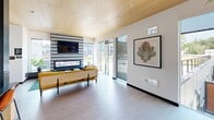 Located at 3795 S Alaska St in Seattle's Columbia City neighborhood, this single-floor community space offers a thoughtfully designed interior complemented by an impressive rooftop amenity area. The interior showcases contemporary architecture highlighted by warm wood-paneled ceilings that lend a refined, natural character throughout. Light hardwood-style flooring flows seamlessly between the living and dining areas, reinforcing a cohesive, open feel.

The living room is a standout space, anchored by a bold geometric-patterned accent wall in black and white, against which a sleek linear fireplace and wall-mounted television are centered. Floor-to-ceiling glass doors and windows flood the room with natural light while providing direct views of the outdoor terrace. The dining area features distinctive patterned wallpaper framed in deep teal, pendant lighting, and a natural wood dining table, creating an inviting atmosphere for gathering. A compact kitchen with light wood cabinetry, stainless steel appliances, and a range hood completes the interior.

The expansive rooftop terrace is a true highlight, offering a putting green, lush landscaped gardens with native plantings, open-air seating with bistro tables, and panoramic views of the surrounding Seattle skyline and neighborhood.