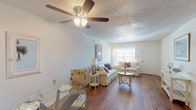 This single-floor space embraces a cohesive coastal theme throughout its open, elongated layout. The living area serves as the centerpiece, featuring warm hardwood flooring that extends across the entire unit, creating a sense of continuity and flow. A textured popcorn ceiling adds depth overhead, while ceiling fans provide both function and character. Natural light filters in through blinded windows, brightening the neutral, light-toned walls that complement the beachy décor accents, including flamingo artwork and starfish-motif pillows.

The living space is generously proportioned, accommodating multiple seating arrangements with ease. A designated dining area sits adjacent, offering a natural transition into the kitchen, which is visible in the background and features white cabinetry and standard appliances. Wall space throughout is well-utilized, with decorative metal palm frond sculptures and coastal artwork enhancing the relaxed, resort-inspired ambiance.

The bathroom is clean and functional, appointed with a white undermount sink set in a light stone-toned vanity, framed wall mirrors, and a shower-tub combination. The overall atmosphere of the space is bright, warm, and inviting, with consistent coastal styling tying each area together seamlessly.
