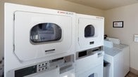 This shared laundry facility is designed to meet the everyday needs of its users with a clean, functional layout. The room houses multiple commercial-grade Maytag stackable washer and dryer units, neatly arranged to maximize the use of available floor space. The stacked configuration allows for a generous number of machines within a compact footprint, ensuring accessibility for multiple users. Each unit is clearly labeled and equipped with coin-operated controls, making them straightforward to operate. The machines are well-maintained and presented in a crisp white finish, contributing to the room's tidy and orderly appearance. The neutral-toned walls and practical design keep the focus on functionality, while the bright interior ensures the space feels open and easy to navigate. Additional top-load washing machines are visible along the side, further expanding the laundry capacity available. Overall, this laundry facility provides a reliable and efficient solution for handling laundry needs, combining quality appliances with a no-frills, user-friendly environment.