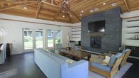 This single-story space is defined by its striking vaulted ceiling, where exposed wooden beams and warm tongue-and-groove wood paneling create an inviting, lodge-inspired atmosphere. A sculptural woven pendant light hangs as a centerpiece, adding an organic, artistic touch to the soaring interior. The main living area is anchored by a floor-to-ceiling slate stone fireplace, which serves as a bold focal point and houses a wall-mounted flat-screen television above its mantle. Comfortable, light grey upholstered sofas and wooden-framed armchairs are arranged around a rectangular wooden coffee table, creating an open and functional seating arrangement ideal for gathering. Large glass panel doors and windows along one wall flood the interior with natural light and provide unobstructed views of well-maintained green lawns and surrounding landscaping. Recessed lighting complements the natural light, ensuring the space is well-lit throughout. The dark flooring grounds the room and contrasts beautifully with the warm wood ceiling and neutral furnishings, contributing to a balanced and welcoming ambiance.