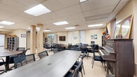 Located at 135 Main St in Poughkeepsie, NY, this single-floor community facility offers a well-organized and functional layout across two distinct areas connected by a corridor. The main gathering space serves as a combined lounge and library, featuring warm beige walls, a drop-panel ceiling with recessed fluorescent lighting, and durable tile flooring with decorative inlay. Multiple round tables with mesh task chairs provide ample seating for group activities or meetings. Bookshelves lined with reading materials flank the walls alongside natural light from tall windows, while a classic upright piano and an American flag add character and a sense of community pride. A leather sofa offers additional casual seating, creating a welcoming, versatile environment. The secondary area functions as a resource and break room, equipped with a commercial double-door refrigerator stocked with food items, a wall-mounted television, shelving units with supplies, and a round table for small group use. Portable room dividers allow for flexible space configuration. A connecting corridor links both areas to additional rooms, including what appears to be a medical or rest space. The overall ambiance is practical, community-oriented, and designed to support a variety of programs and services.