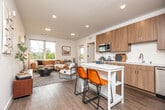 This contemporary single-floor residence offers a thoughtfully designed open-concept layout that seamlessly connects the kitchen, dining, and living areas. The kitchen is a standout feature, outfitted with warm wood-grain cabinetry extending to the ceiling, white quartz countertops, a subway tile backsplash, and a full suite of stainless steel appliances including a range, microwave, dishwasher, and French door refrigerator. A compact island with seating provides a convenient casual dining option. The living area is bathed in natural light from large windows and a glass door, offering outdoor views and creating an airy, welcoming atmosphere. Wood-look flooring runs consistently throughout the main areas, anchoring the modern aesthetic. Two well-proportioned bedrooms each feature windows that invite natural light, clean white walls, and easy access to the bathrooms. Both bathrooms are finished with wood-tone vanities, white quartz countertops, and full soaking tub and shower combinations. The unit also includes an in-unit washer and dryer, adding a practical convenience. The overall design palette of whites, warm wood tones, and brushed metal finishes creates a cohesive, contemporary ambiance throughout the space.
