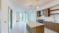 This contemporary 2-bedroom, 1-bathroom unit offers a thoughtfully designed open-concept layout that seamlessly connects the kitchen, dining, and living areas. The kitchen is a standout feature, showcasing warm wood-tone flat-panel cabinetry, a white subway tile backsplash, a large waterfall-edge island with pendant lighting overhead, and a full suite of stainless steel appliances including a range, dishwasher, built-in microwave, and french door refrigerator. Light-toned hardwood-style flooring runs throughout the main living areas, complementing the bright, neutral palette of the space. The living room is bathed in natural light, courtesy of expansive floor-to-ceiling sliding glass doors that open to a private balcony overlooking the surrounding cityscape. Large-format light tile flooring continues into the bedrooms, which are well-proportioned and serene, including a generously sized primary bedroom. The bathroom features a floating wood vanity with a white quartz countertop, an undermount sink, and an illuminated frameless mirror, delivering a clean and contemporary aesthetic. A dedicated closet and clean hallways with modern flat-panel doors complete the functional layout of this well-appointed urban residence.