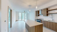 This contemporary 2-bedroom, 1-bathroom unit offers a thoughtfully designed open-concept layout that seamlessly connects the kitchen, dining, and living areas. The kitchen is a standout feature, showcasing warm wood-tone flat-panel cabinetry, a white subway tile backsplash, a large waterfall-edge island with pendant lighting overhead, and a full suite of stainless steel appliances including a range, dishwasher, built-in microwave, and french door refrigerator. Light-toned hardwood-style flooring runs throughout the main living areas, complementing the bright, neutral palette of the space. The living room is bathed in natural light, courtesy of expansive floor-to-ceiling sliding glass doors that open to a private balcony overlooking the surrounding cityscape. Large-format light tile flooring continues into the bedrooms, which are well-proportioned and serene, including a generously sized primary bedroom. The bathroom features a floating wood vanity with a white quartz countertop, an undermount sink, and an illuminated frameless mirror, delivering a clean and contemporary aesthetic. A dedicated closet and clean hallways with modern flat-panel doors complete the functional layout of this well-appointed urban residence.